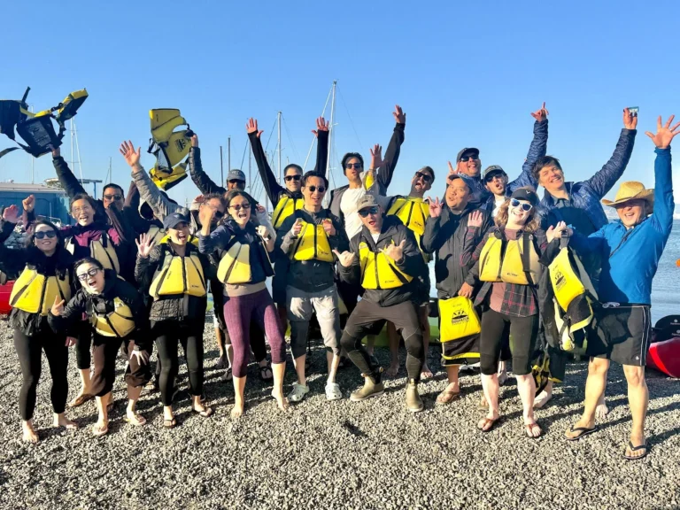 A large group on the beach at Crane Cove in life jackets, arms raised, the gantry crane behind them.