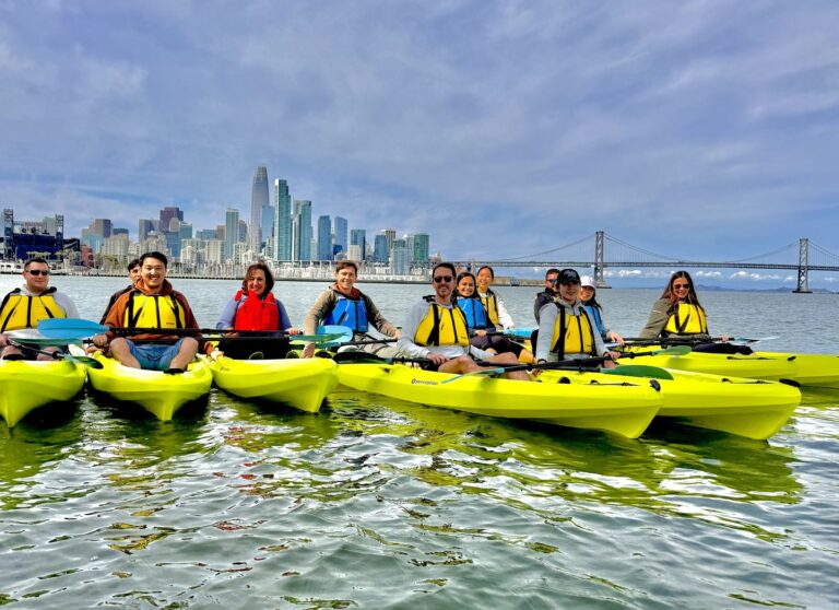 A group in yellow kayaks rafted together with the city skyline behind them.