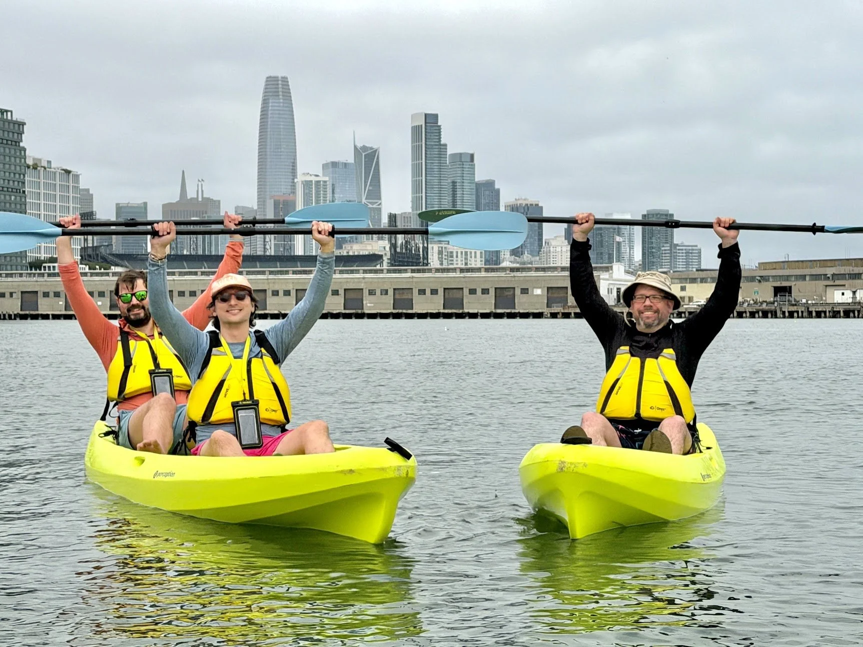Three paddlers in yellow kayaks raising their paddles overhead with the downtown skyline behind.