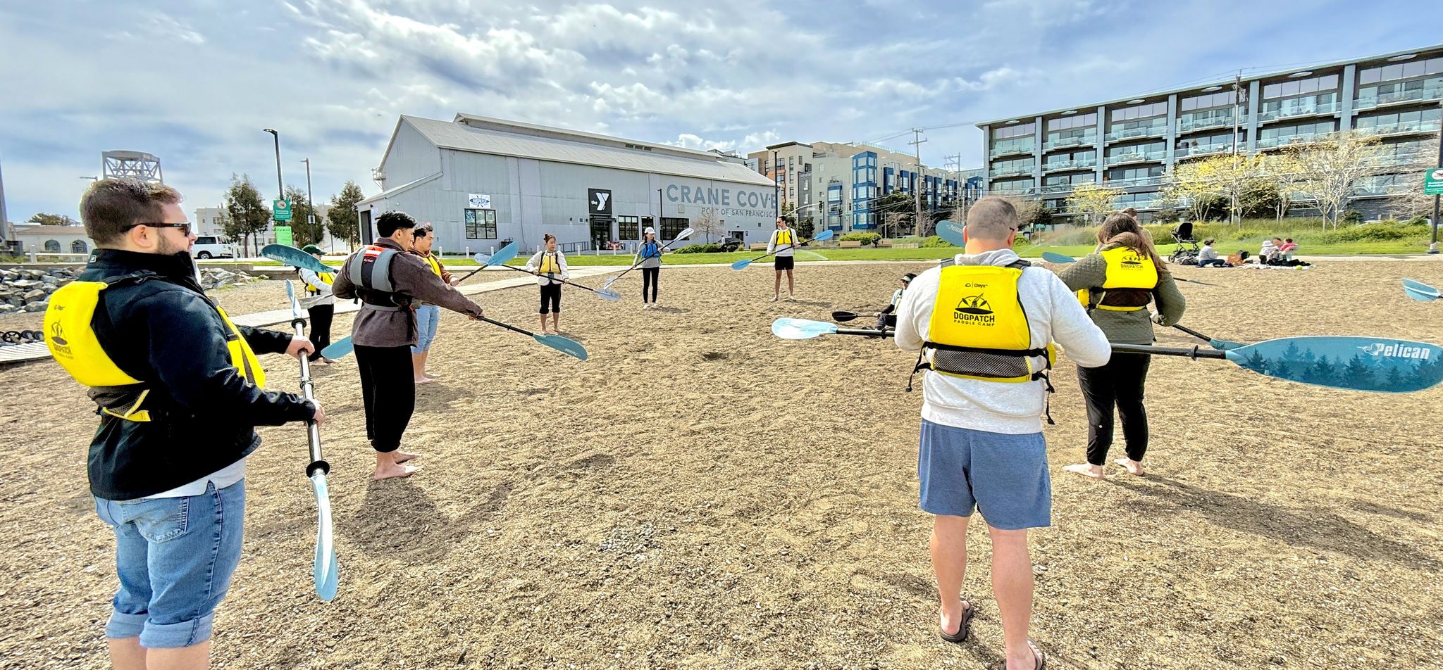 A paddle briefing on the beach at Crane Cove Park: a circle of people in yellow life vests holding kayak paddles out in front of them, Building 49 and the Dogpatch shoreline behind them