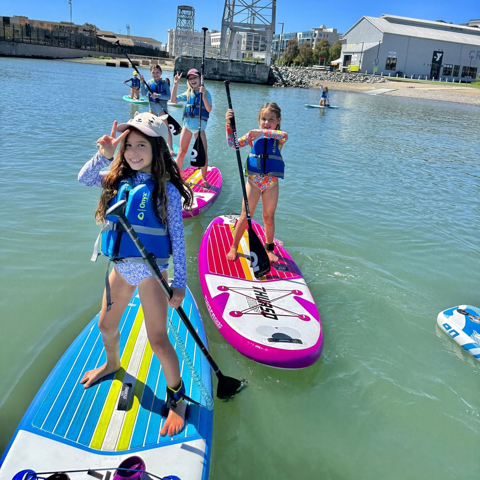 Young campers on stand-up paddle boards in the protected water at Crane Cove Park, with Building 49 and the Pier 70 gantry behind them