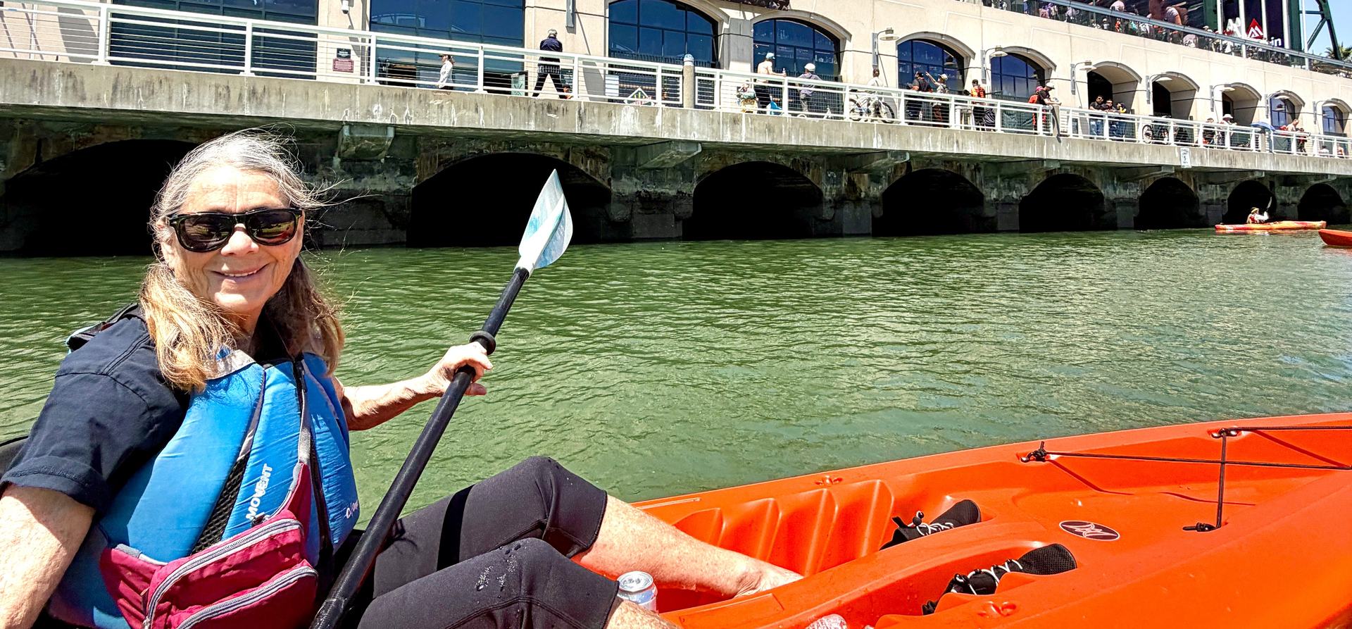 A paddler in an orange Dogpatch tandem kayak on McCovey Cove, with the arcade at Oracle Park and the crowd along the waterfront rail behind her