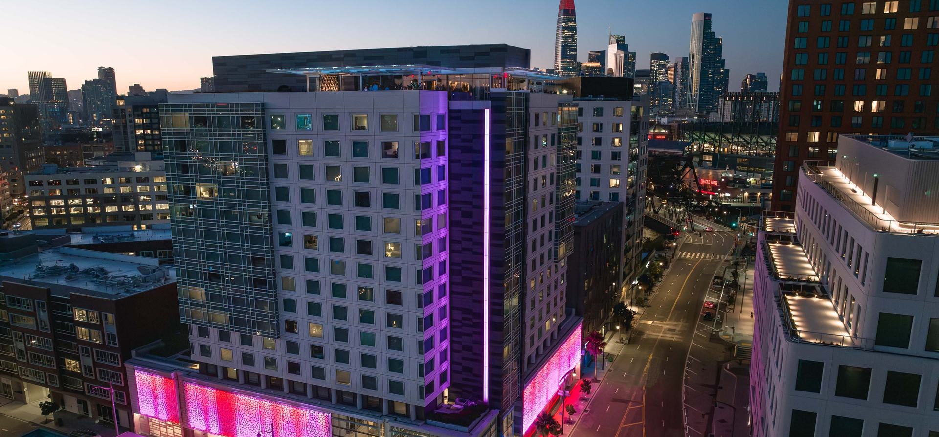 LUMA Hotel San Francisco lit magenta and violet at dusk, with the downtown skyline and Oracle Park behind it