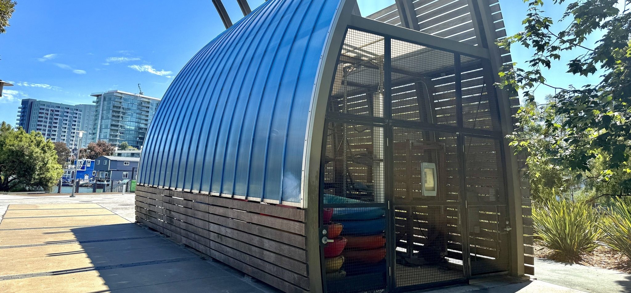 The Mission Creek Boathouse at 401 Berry Street, its curved blue roof in profile, kayaks racked behind the mesh gate, and the Mission Creek houseboats and Mission Bay towers behind it