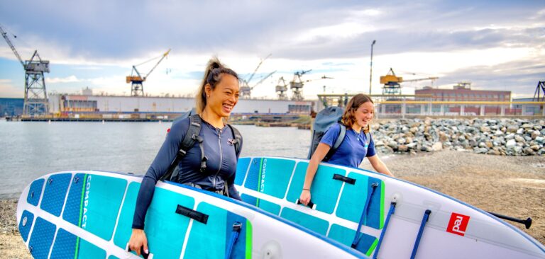 Two paddlers carrying inflatable paddle boards down the beach at Crane Cove Park, with the Pier 70 shipyard cranes behind them