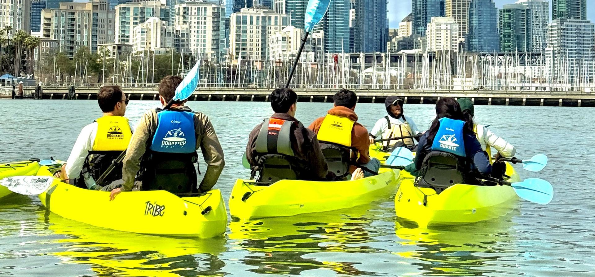 A group of paddlers in Dogpatch buoyancy aids, seen from behind in yellow tandem kayaks, with the San Francisco skyline and a marina behind them